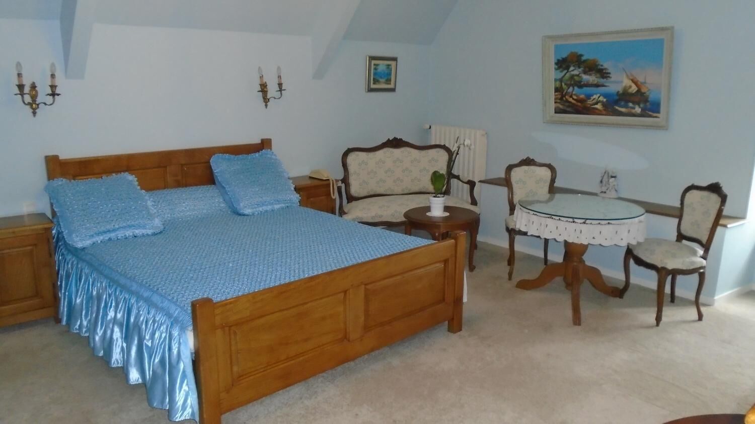 Un dormitorio con una cama de madera, una mesita redonda pequeña, dos sillas, una mesita de noche y una lámpara de pared.