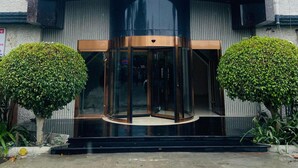 Un edificio moderno con fachada curva, puerta giratoria y dos arbustos verdes simétricos.