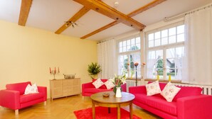 A living room with a red sofa set, a wooden coffee table, and a red rug. There is a window with curtains and a view of greenery outside. - Mueritz Landhausperle (Röbel/Müritz)