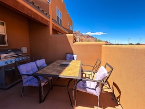 An outdoor patio with a grill, table, and chairs, set against a building with a textured wall and a view of a mountain.