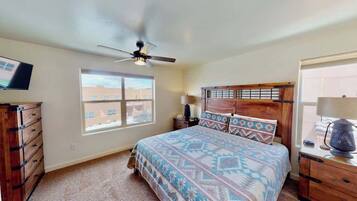 A bedroom with a bed, a ceiling fan, a nightstand, a lamp, a dresser, and a window with a view of buildings.