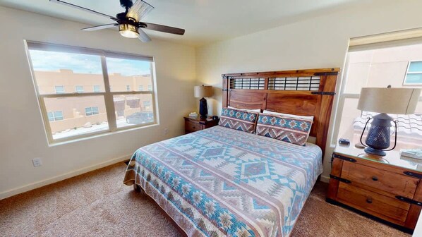 Une chambre à coucher comprenant un lit, une table de chevet, une commode, un ventilateur de plafond et une fenêtre donnant sur des immeubles.