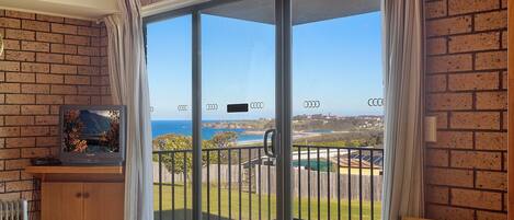 A room with a sliding glass door leading to a balcony with a view of the sea and a distant landscape.
