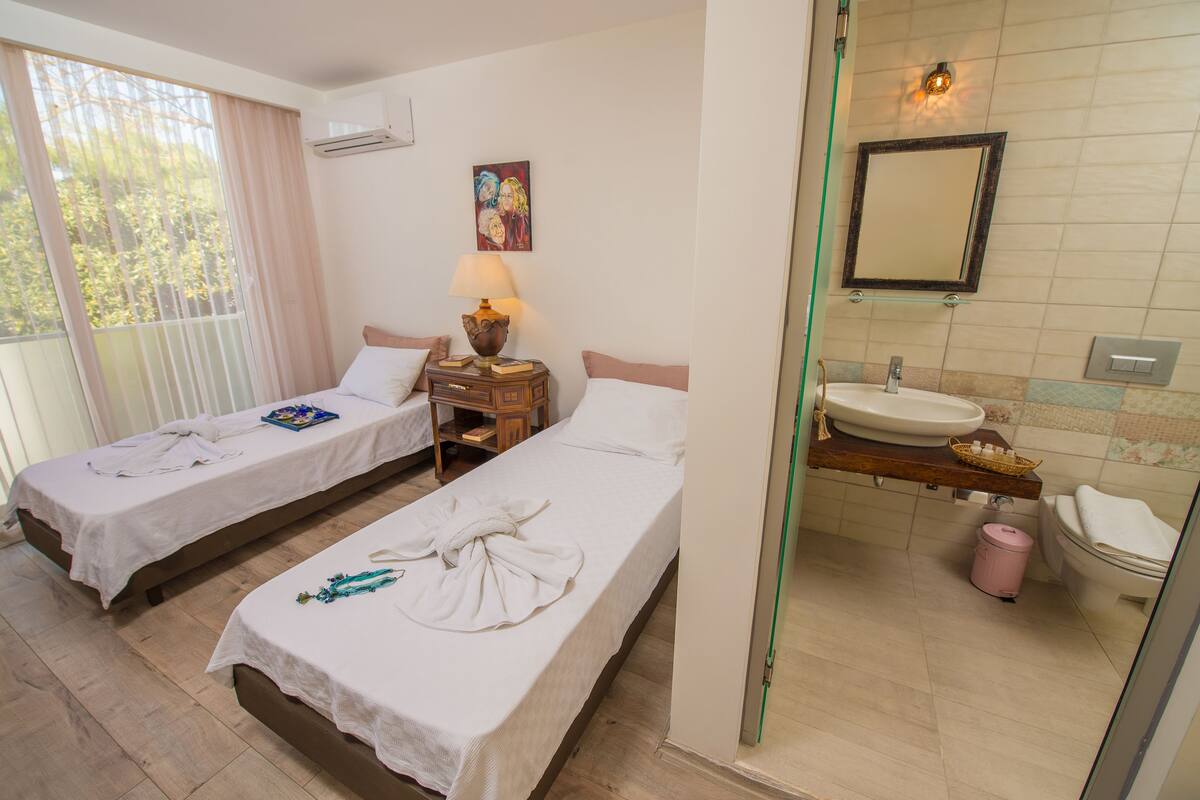 Ein Hotelzimmer mit zwei Betten, einem Badezimmer inklusive Waschbecken und Spiegel sowie einem Fenster mit Vorhängen.