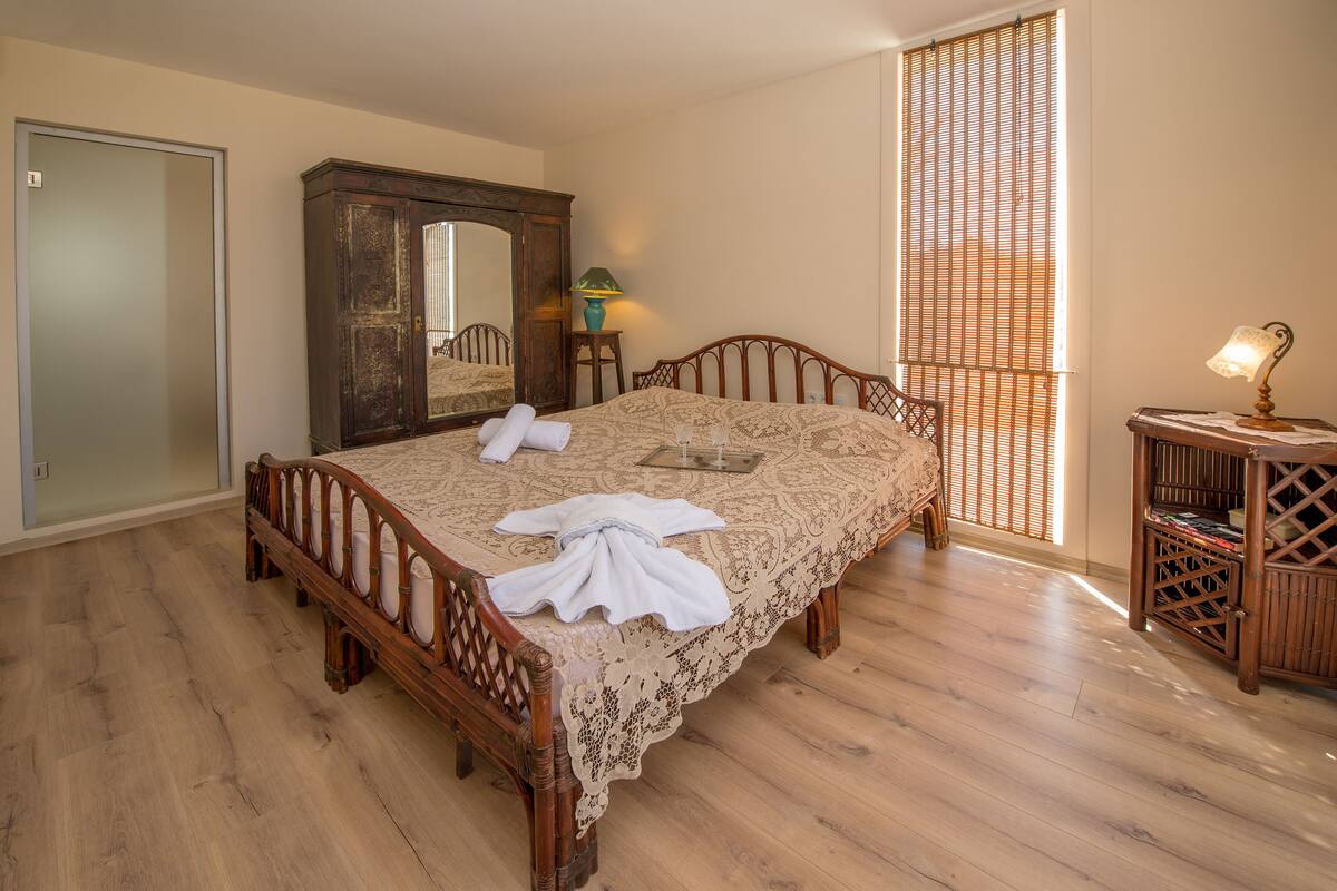 Ein ordentlich bezogenes Bett mit Häkeldecke, ein Holzschrank, ein Spiegel und ein Fenster mit Jalousien.