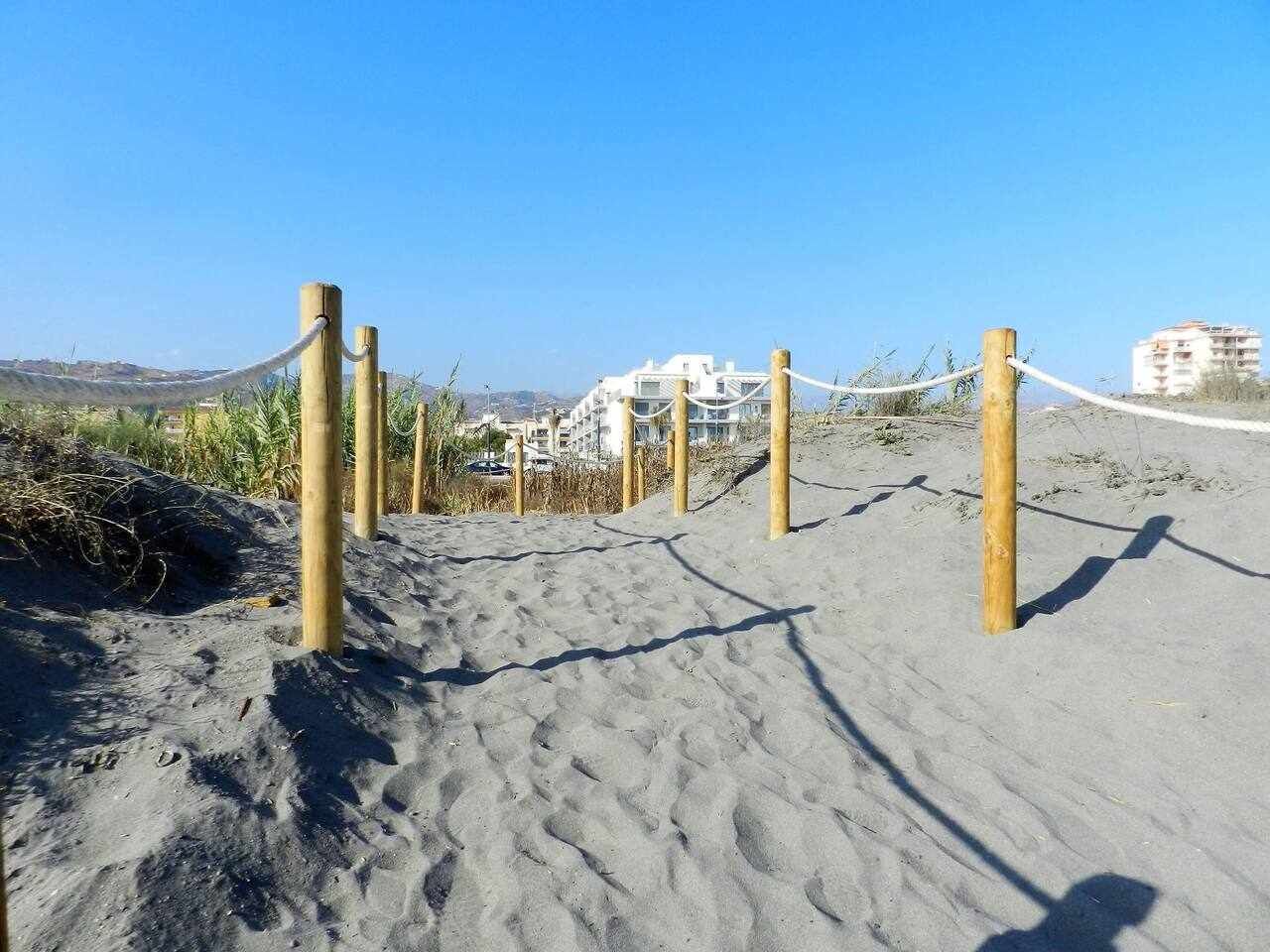 A sandy beach path with wooden posts and ropes, leading to buildings in the background.