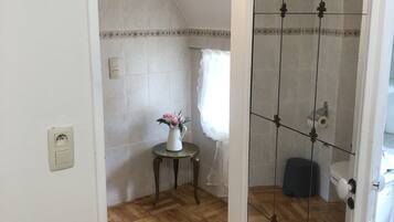 Una habitación con suelo de madera en espiga, pared blanca con un detalle decorativo, una mesita con un florero y una zona de ducha con puerta de vidrio.