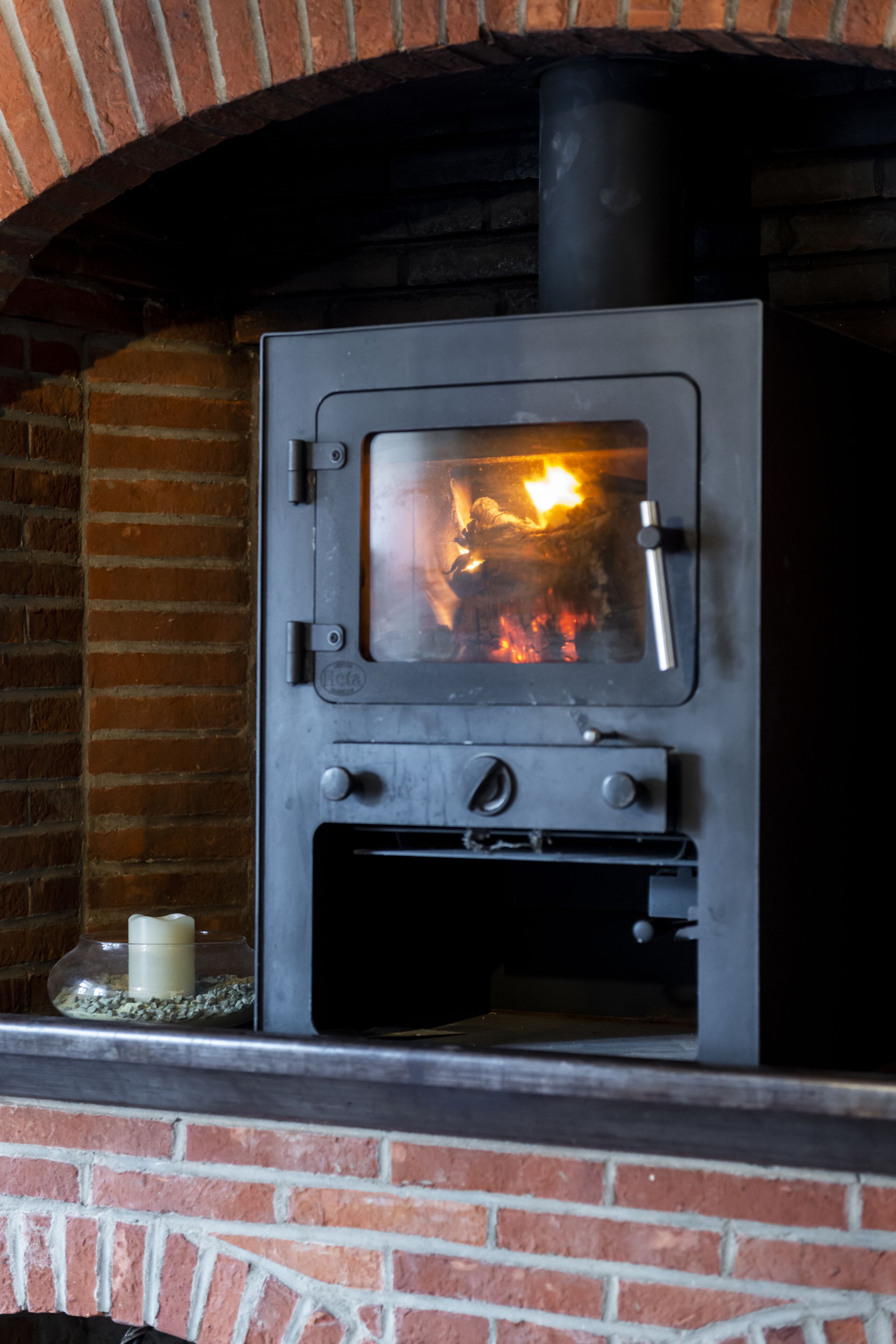 A wood-burning stove with a glass door, a control panel, and a brick chimney.