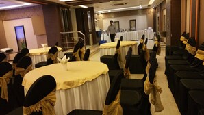 A banquet hall with round tables covered in white tablecloths, black chairs with gold sashes, and a person walking in the background. - HOTEL ACCORD (Ranchi)