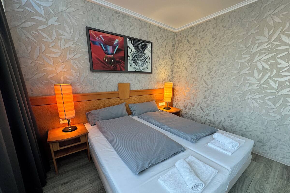 A neatly made bed with pillows and a folded towel, two bedside lamps, a framed artwork above the bed, and a patterned wallpaper.