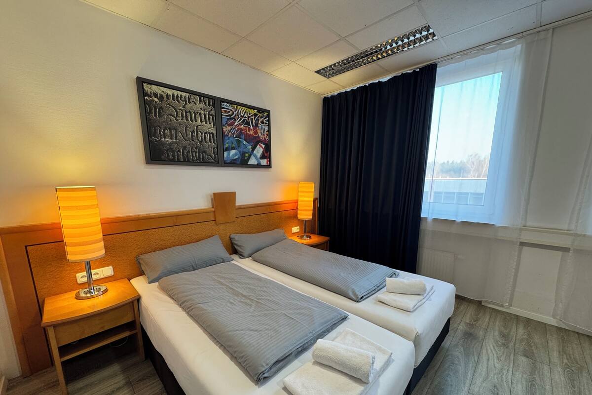 A hotel room with two beds, a headboard, bedside lamps, a window with curtains, and artwork on the wall.