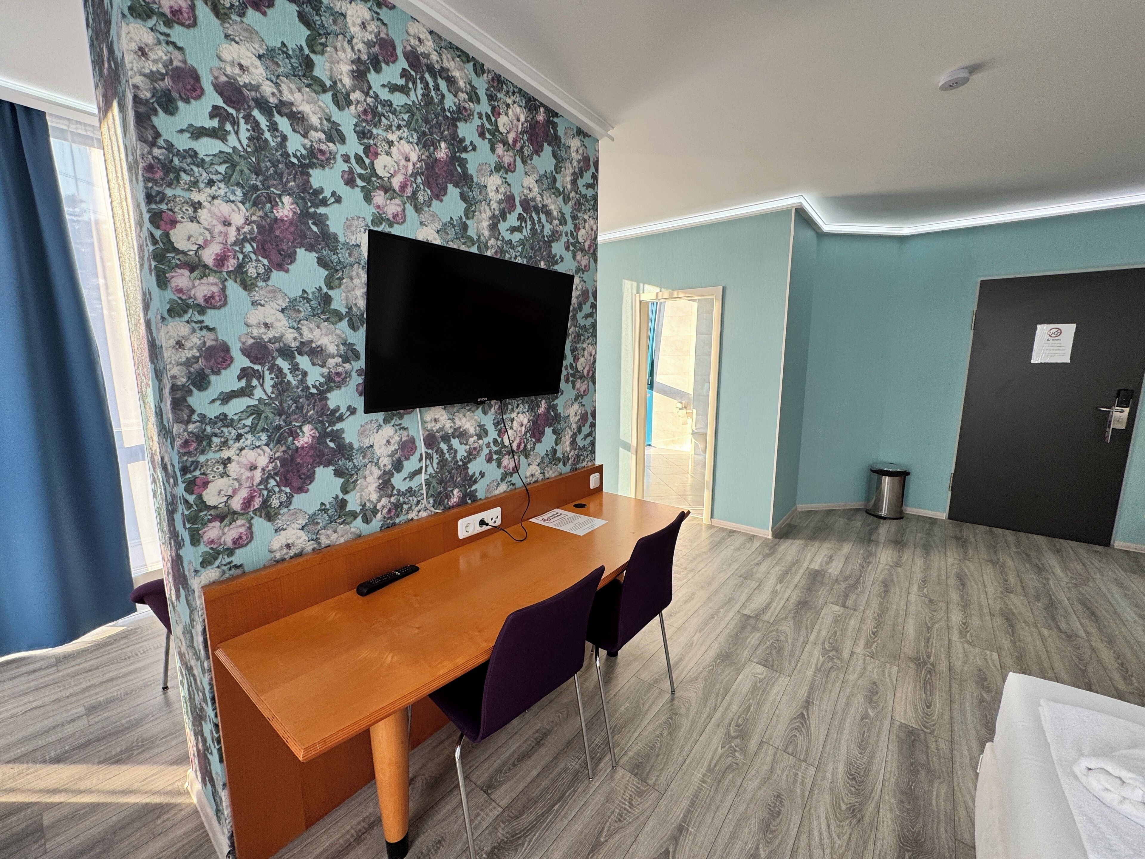 A hotel room with a wooden dining table, purple chairs, a flat-screen TV mounted on a floral wallpapered wall, and a door with a trash bin beside it.