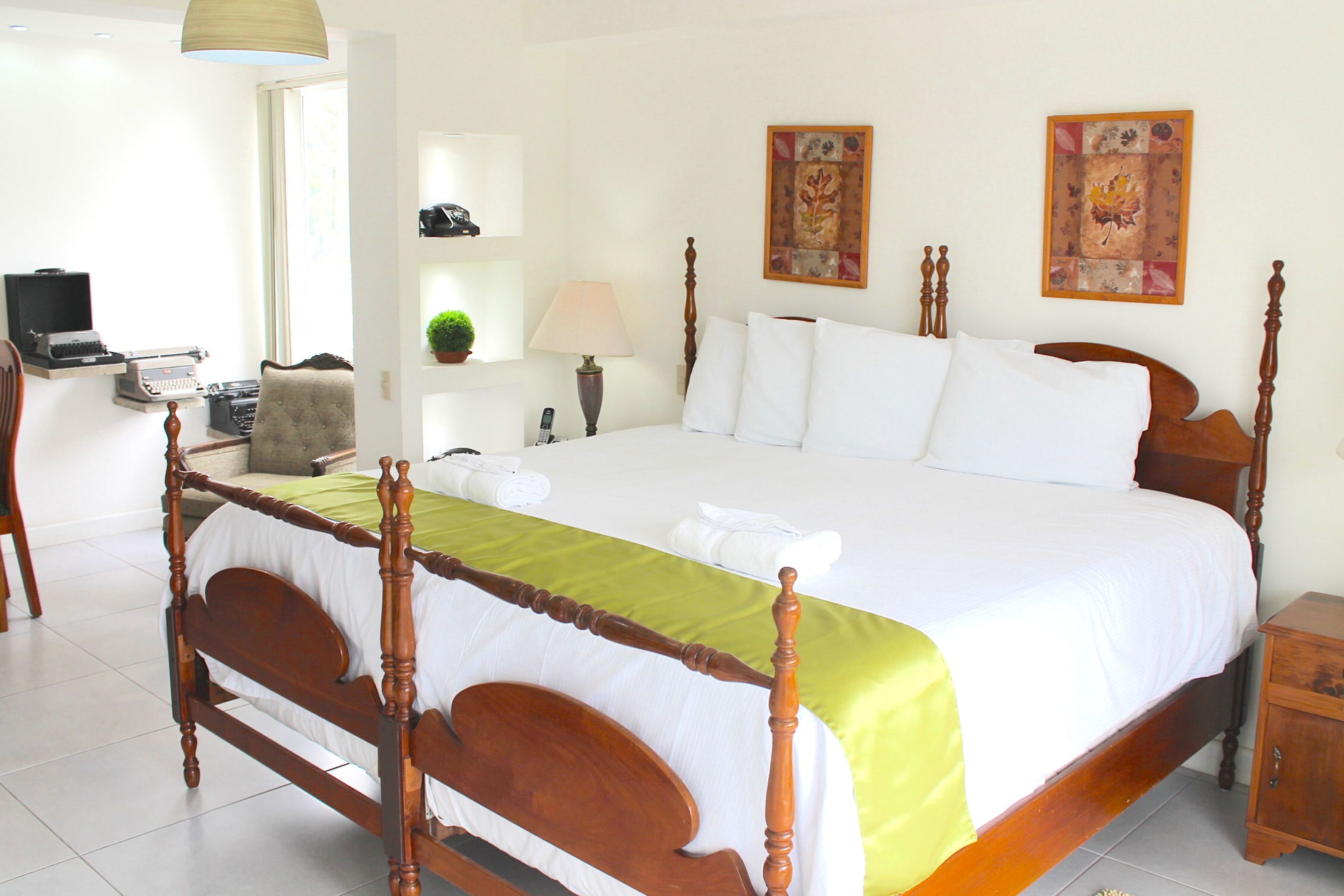 Una cama bien hecha con estructura de madera, colcha verde y almohadas blancas. Se observa una mesita de noche con una lámpara y un objeto decorativo en la pared.