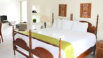 Una cama bien hecha con estructura de madera, colcha verde y almohadas blancas. Se observa una mesita de noche con una lámpara y un objeto decorativo en la pared.