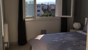 A bedroom with a large bed, a window with curtains, a bedside table with a lamp, and a view of buildings and mountains outside. - Furnished apartment T2 St Martin D Héres (Saint-Martin-d'Hères)