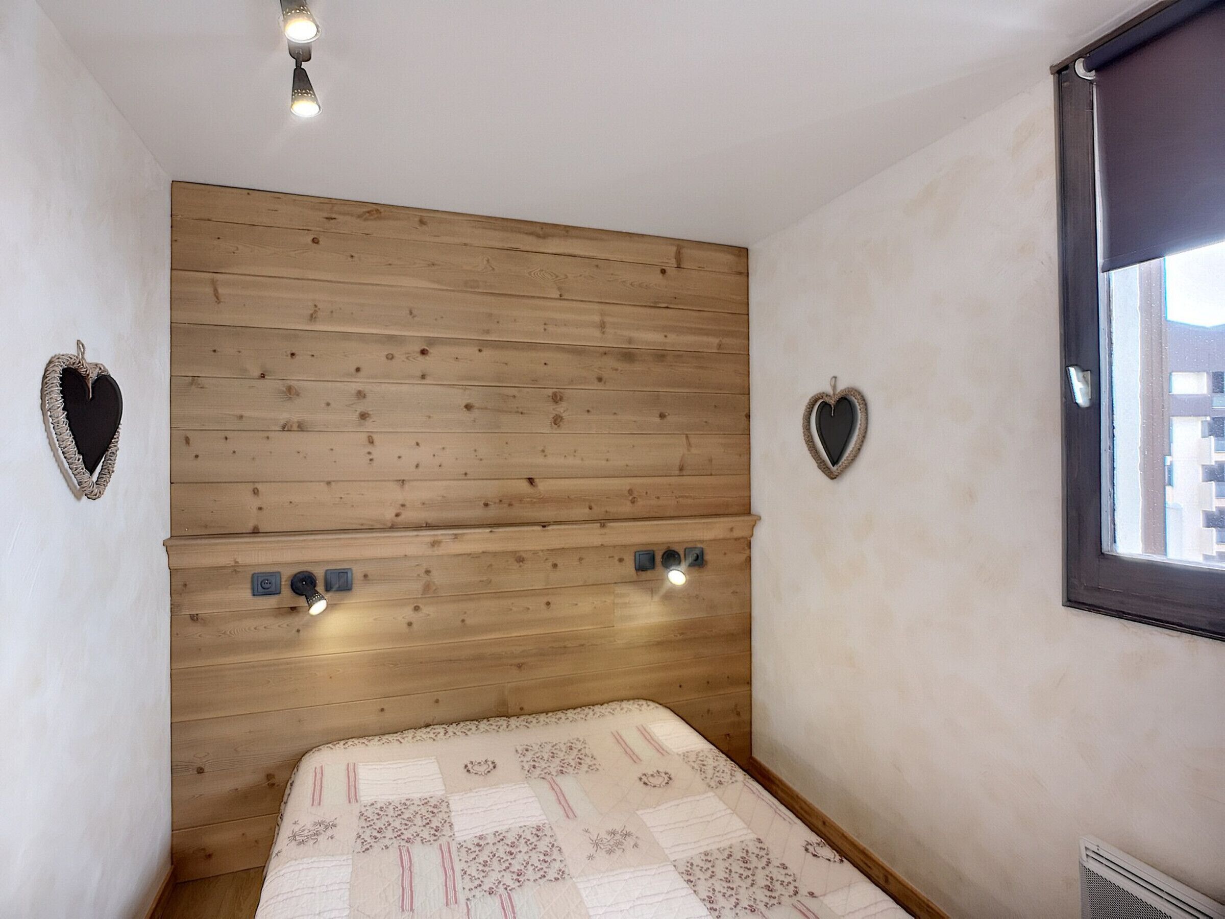 Een slaapkamer met een houten hoofdbord, twee hartvormige decoraties aan de muur, een bed met een bedsprei met patroon, en een raam met uitzicht op gebouwen.