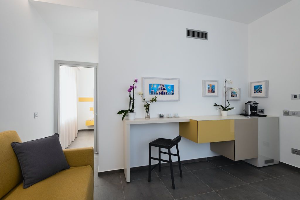 Eine moderne Küche mit weißer Arbeitsplatte, einem gelben Schrank und einem schwarzen Hocker. An der Wand hängen Bilder in Rahmen und auf der Arbeitsplatte steht ein Blumentopf.