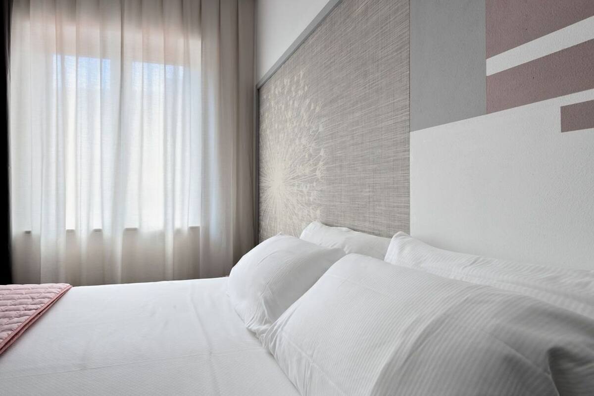 A hotel room with a large bed, white linens, a pink throw, and sheer curtains.