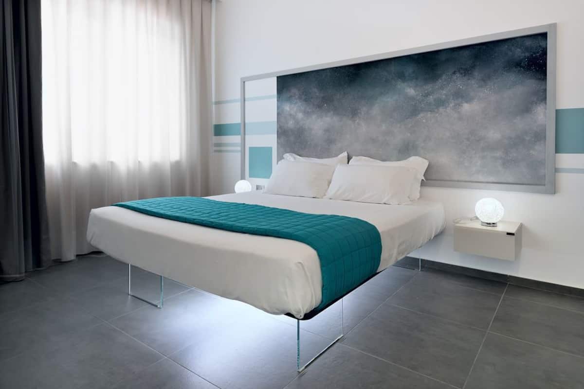 A modern hotel room with a large bed, a nightstand with a lamp, and a large abstract painting on the wall.