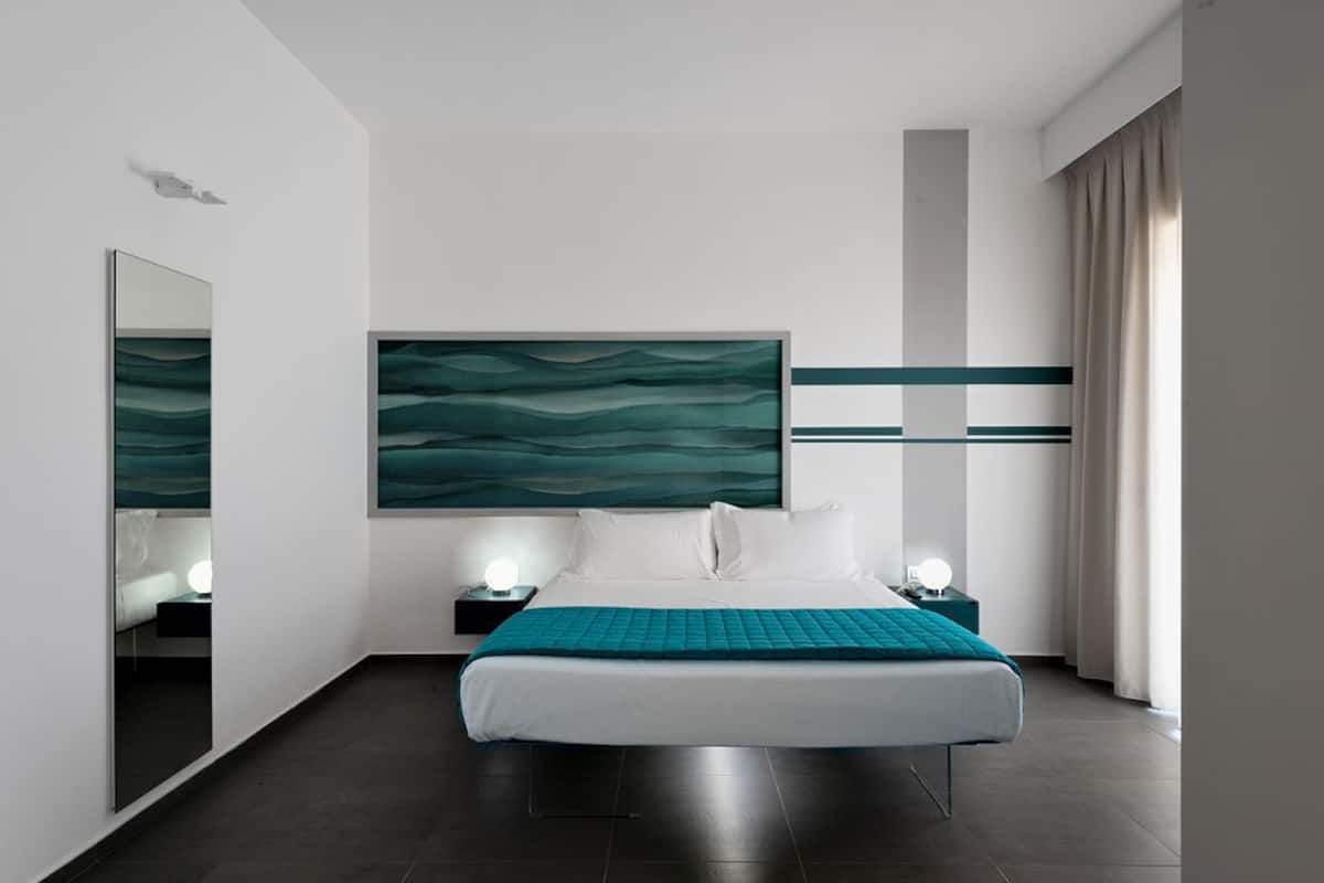 A modern hotel room with a large bed, two bedside tables with lamps, a large abstract painting on the wall, and a view of the city through the window.