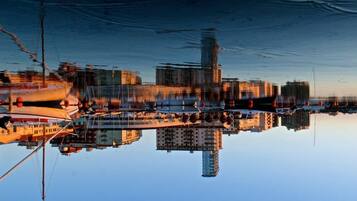 A waterfront cityscape with buildings reflected in calm water, boats docked along the shore, and a clear sky.