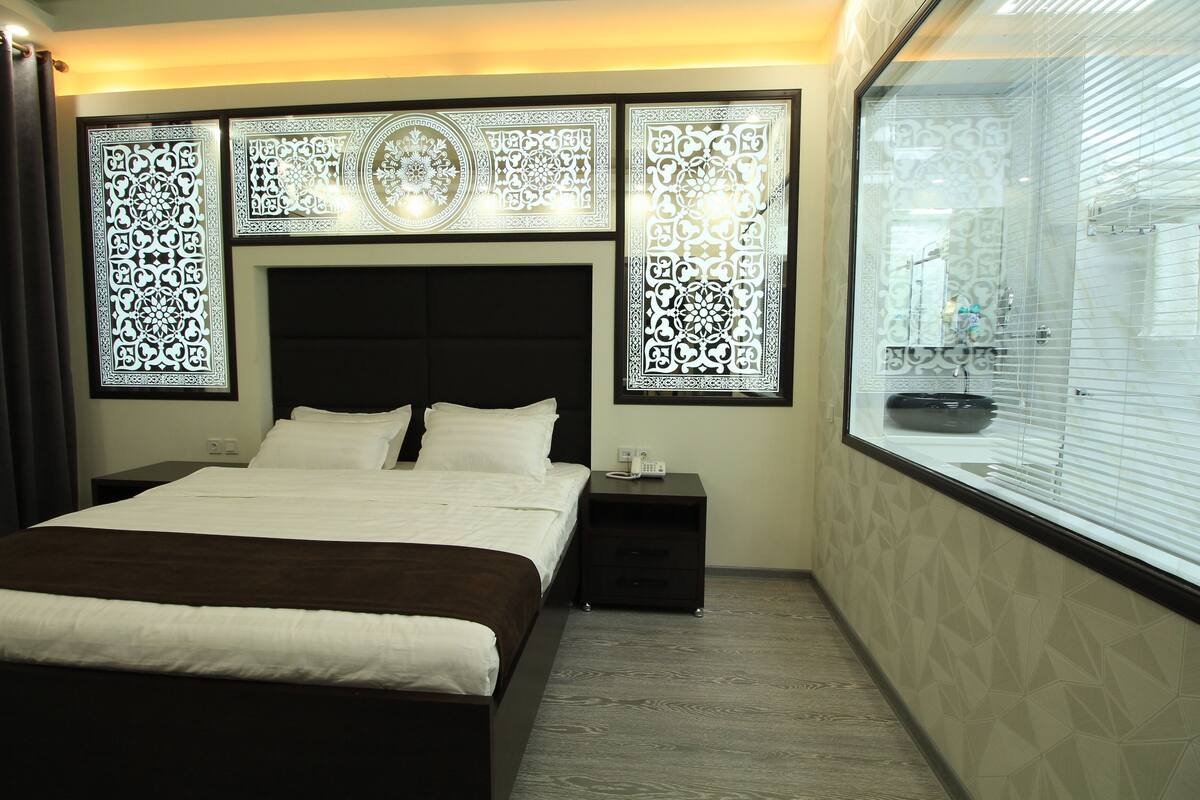 A hotel room with a large bed, a nightstand, a decorative wall panel, and a window with blinds.