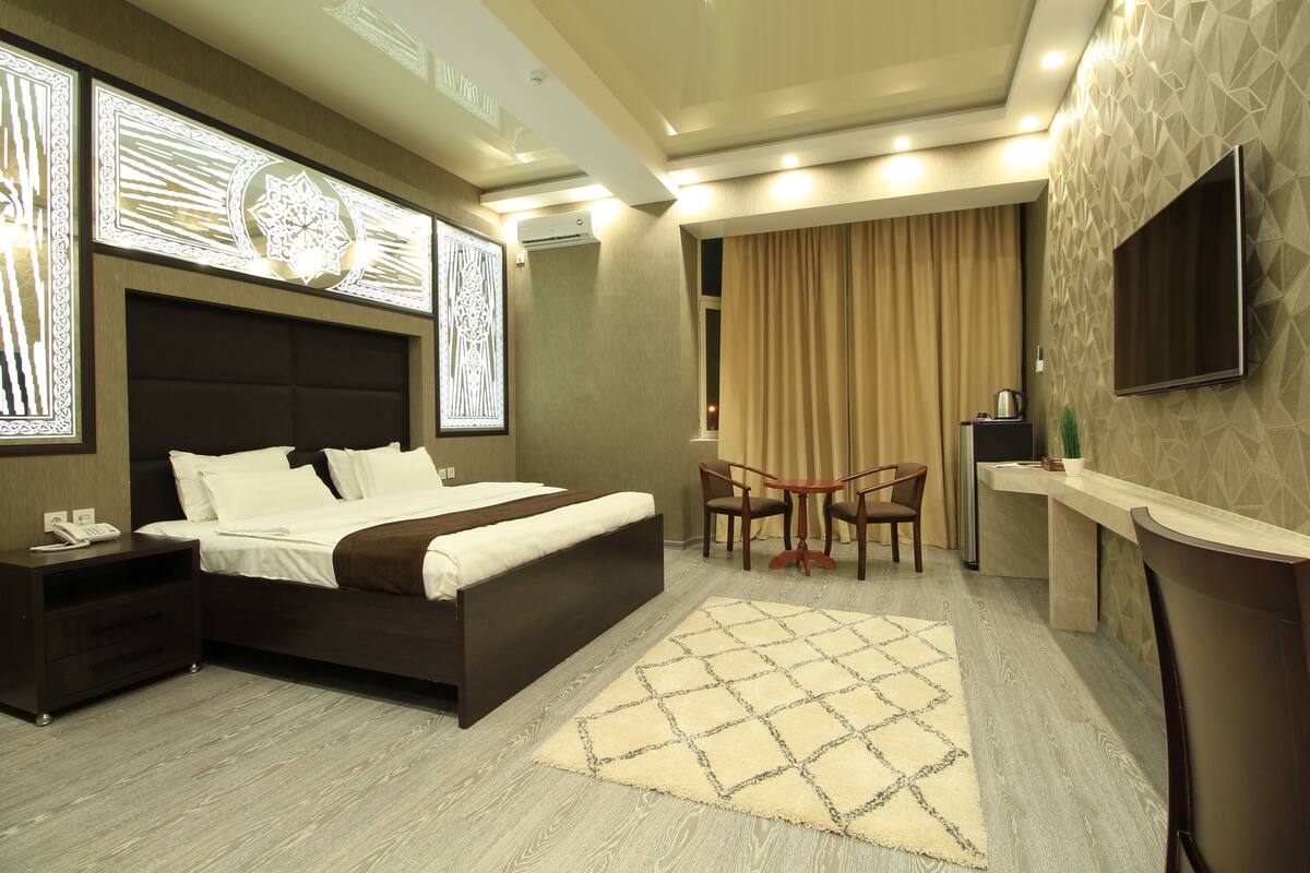 A modern hotel room with a large bed, bedside tables, a desk, a chair, and a TV mounted on the wall.
