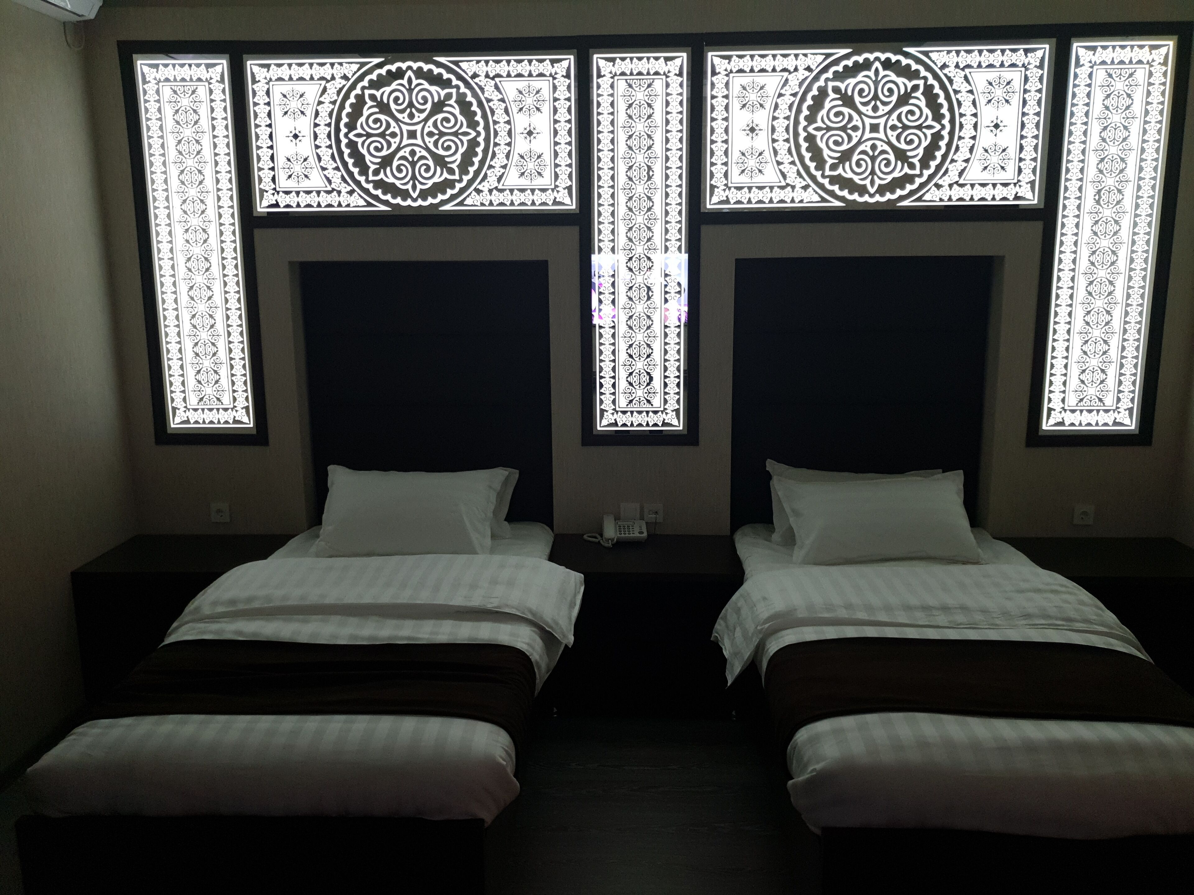 A hotel room with two beds, each with a dark headboard and white bedding. There are decorative window patterns above the beds.