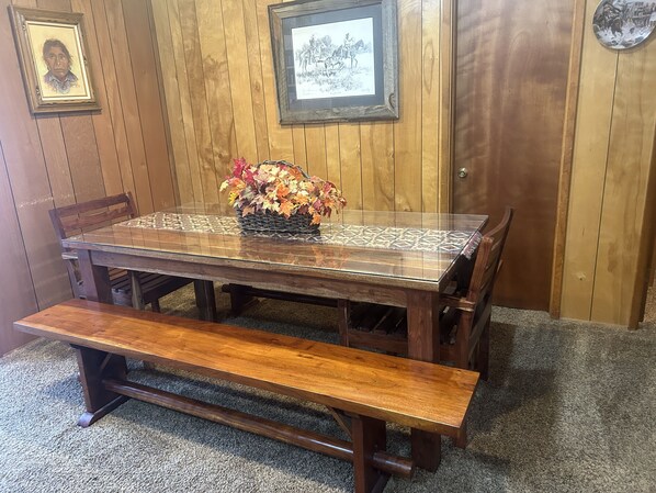Una mesa de comedor de madera con cubierta de vidrio, rodeada de sillas. En frente de la mesa hay un banco.