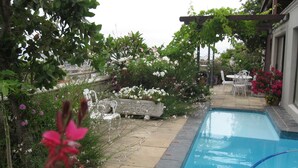 A swimming pool with a stone border, surrounded by a garden with various plants and flowers, and a pergola with a table and chairs underneath. - 123 Ocean View Drive Apartment (Cape Town)
