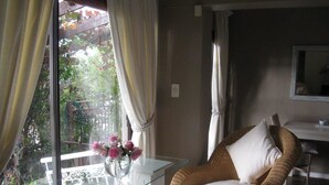 A room with a wicker chair, a glass table with a vase of flowers, and a view of greenery through a window with curtains. - 123 Ocean View Drive Apartment (Cape Town)