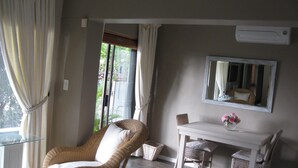 A room with a wicker chair, a small table with a vase of flowers, a mirror on the wall, and a window with curtains. - 123 Ocean View Drive Apartment (Cape Town)