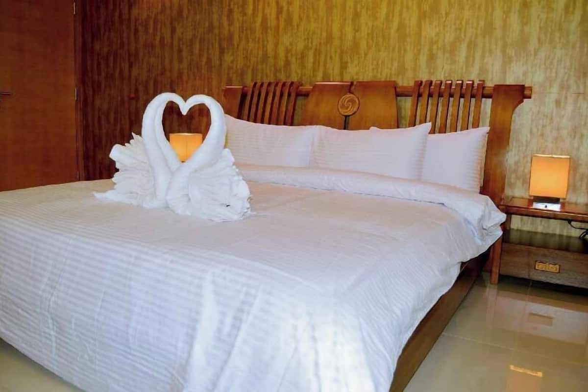 Una cama bien hecha con un adorno con forma de corazón en la colcha, un cabecero de madera y una mesita de noche con una lámpara.