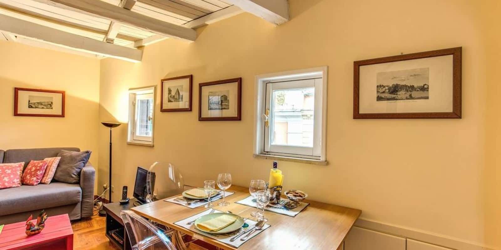 A dining area with a wooden table set for two, framed pictures on the wall, and a window providing natural light.
