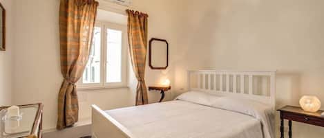 A bedroom with a bed, a bedside table with a lamp, a window with curtains, and a wall-mounted mirror.