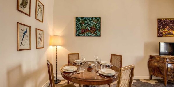 A dining area with a round table set for four, surrounded by wicker chairs. There are framed artworks on the walls, a floor lamp, and a television on a wooden cabinet. - Principe Amedeo 2 (Rome)