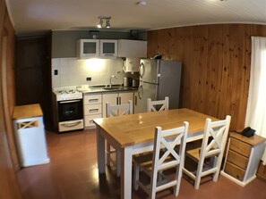 A compact kitchen with white cabinets, a refrigerator, and a stove. There is a wooden dining table with chairs. - Cabañas Altue (El Calafate)