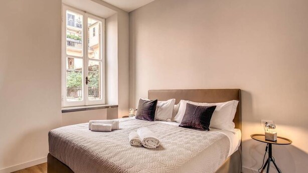Una cama bien hecha con edredón blanco y almohadas, una mesita de noche con lámpara, y una ventana con vistas a edificios y vegetación.