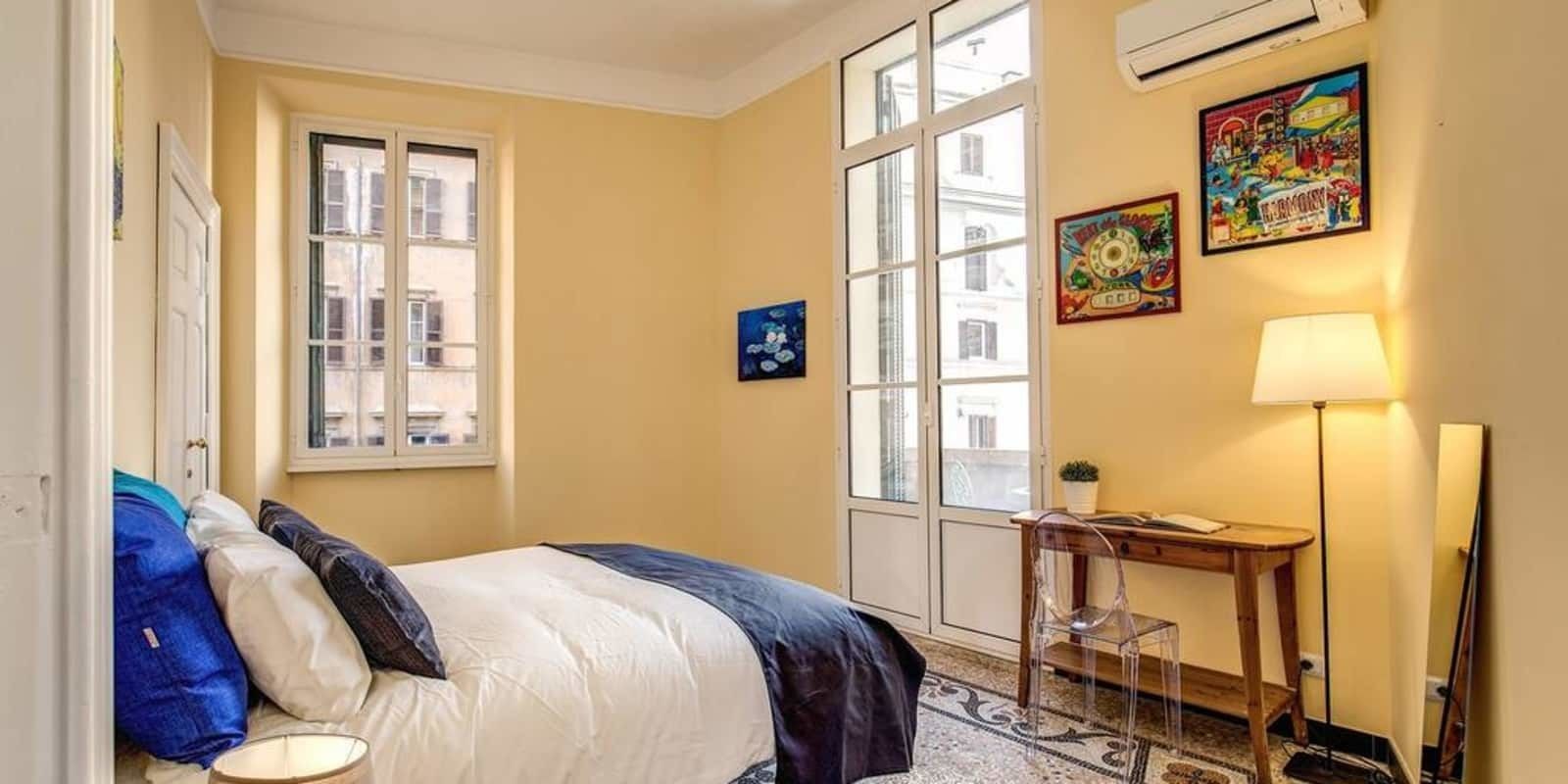 A hotel room with a bed, a desk, a chair, a lamp, and a window.