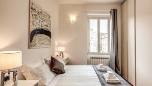 A hotel room with a bed, a nightstand, a lamp, a window with curtains, and a radiator. - Attic terrace with St Peter's Dome View (Rome)
