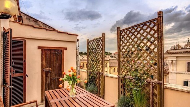 A balcony with a wooden table and a vase of flowers, a wooden gate, and a view of buildings in the background. - Attic terrace with St Peter's Dome View (Rome)