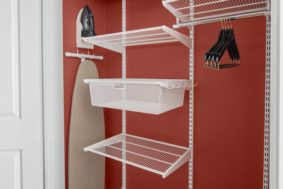 Una pared roja con estantes blancos que sostienen un planchador, una cesta y zapatos negros.