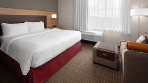 Habitación de hotel con una cama grande, un pequeño sofá, una mesita de noche, una lámpara y una ventana con cortinas translúcidas.