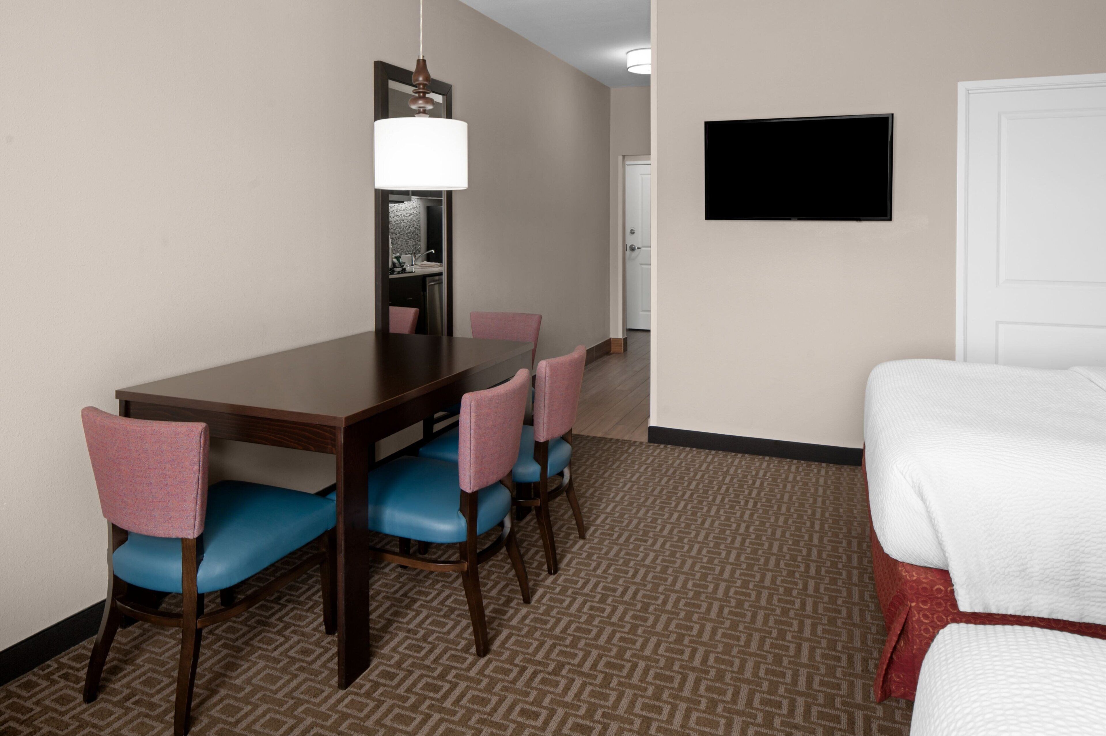 A hotel room with a wooden dining table, blue chairs, a large mirror, a flat-screen TV, and a bed with white bedding.