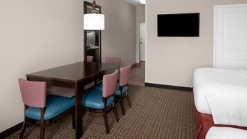A hotel room with a wooden dining table, blue chairs, a large mirror, a flat-screen TV, and a bed with white bedding.