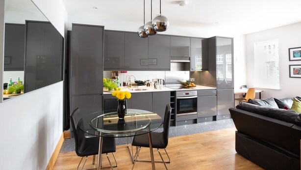 A modern kitchen with dark cabinets, a glass dining table, and a black sofa. - HomefromHolme Alban House (St Albans)