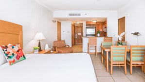A hotel room with a bed, a chair, a small table, a lamp, and a view of the kitchen and dining area. - Condado Lagoon Hospitality (San Juan)