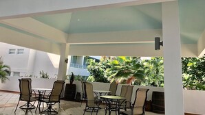An outdoor patio area with a thatched roof, surrounded by potted plants and a view of a building in the background. - Condado Lagoon Hospitality (San Juan)