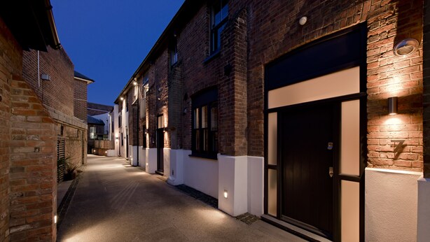 A narrow alleyway with brick buildings, illuminated by wall-mounted lights, leading to a dark entrance. - HomefromHolme St Peters Mews (St Albans)