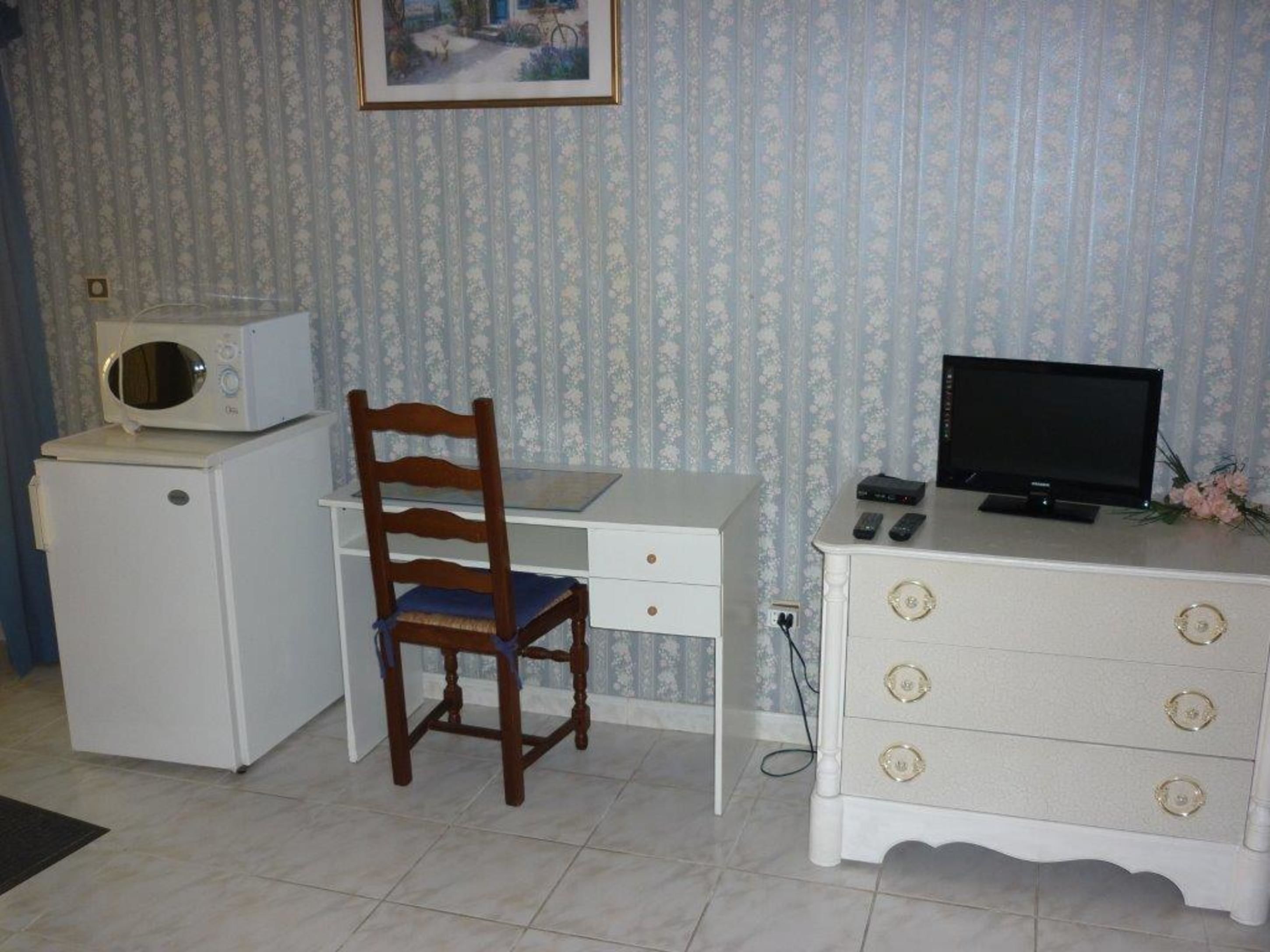 A compact room with a white desk, a wooden chair, a small white cabinet with drawers, a television, and a microwave on a small white appliance.